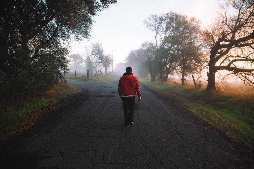 A man wearing a red jacket walking away doing a rural road on a foggy day